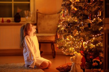 Gecelik giyip güzel bir Noel ağacının yanında oturan tatlı küçük kız. Noel ve Yeni Yıl konsepti. Sihrin atmosferi, ışıklar, ışıltılar. Mutlu çocuk hediyeyle ağaca bakıyor.