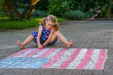 Gözlüklü, yıldız ve çizgili kıyafetler giymiş küçük bir kız, Amerikan bayrağının büyük bir tebeşir çiziminin yanında asfalt bir yüzeye oturur. Mutlu çocuk asfalta Amerikan bayrağı çiziyor.