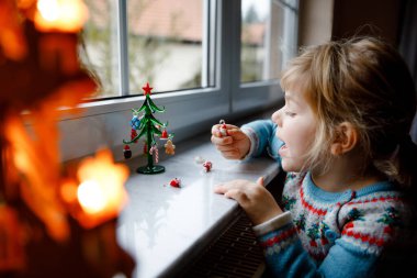 Küçük kız pencerenin kenarında oturuyor ve küçük cam Noel ağacını küçük Noel oyuncaklarıyla süslüyor. Mutlu sağlıklı çocuk geleneksel aile bayramını kutluyor. Sevimli bebek