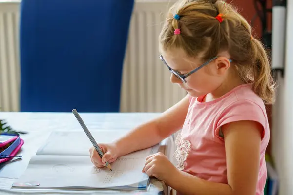 stock image A young girl with glasses, sitting at a table, is focused on writing in her workbook. Child practicing writing numbers with a pencil, engaged in learning. Elementary school, education