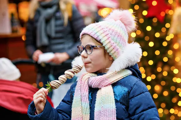 Geleneksel Alman Noel pazarında beyaz çikolata kaplı çilek yiyen küçük tatlı kız. Karlı bir günde Almanya 'da geleneksel aile pazarında mutlu bir çocuk.