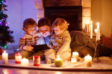 Genç anne ve Noel zamanında bir şöminenin evde oturan iki küçük çocuğu. Tablet ile oynayan çocuklar çocuklar. Üç sarılma aile. Mutlu kadın ve sevimli oğlu, İkizler küçük çocuklar.