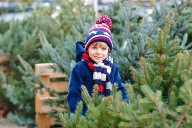 Noel ağacı tutan komik küçük çocuk boy gülümseyerek. Seçme ve xmas ağacı açık dükkanda satın Kış moda giysileri mutlu bir çocuk. Aile, geleneği, kutlama.