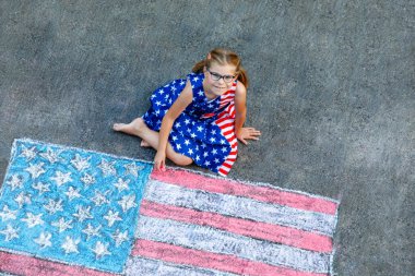 Little girl with glasses, dressed in a stars and stripes outfit, sits on a paved surface beside a large chalk drawing of the American flag. Happy child painting USA flag on asphalt. clipart