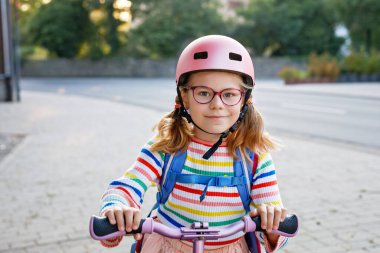 Scooter sürerek okula giden mutlu küçük kız. Büyük bir çantayla şehirde gezen ilkokul çocuğu. Pembe kasklı çocuk. Okula güvenli yol ve çocuk konsepti için hareket.