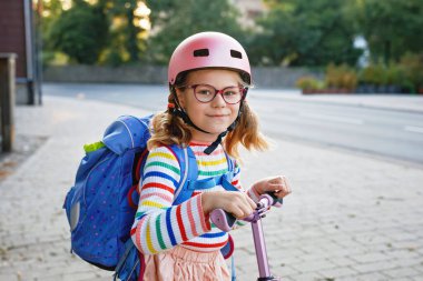Scooter sürerek okula giden mutlu küçük kız. Büyük bir çantayla şehirde gezen ilkokul çocuğu. Pembe kasklı çocuk. Okula güvenli yol ve çocuk konsepti için hareket.