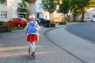 Little happy girl on way to school by pushing scooter. Elementary school child riding in the city, with big satchel. Kid with pink helmet. Safe route to school and movement for children concept. clipart