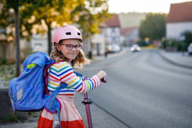 Scooter sürerek okula giden mutlu küçük kız. Büyük bir çantayla şehirde gezen ilkokul çocuğu. Pembe kasklı çocuk. Okula güvenli yol ve çocuk konsepti için hareket.