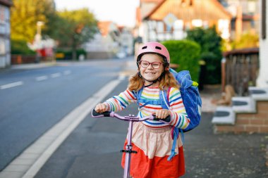 Scooter sürerek okula giden mutlu küçük kız. Büyük bir çantayla şehirde gezen ilkokul çocuğu. Pembe kasklı çocuk. Okula güvenli yol ve çocuk konsepti için hareket.