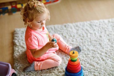 Mutlu küçük kız halıya oturmuş, renkli tahta halkalar ve oyuncak kamyonla oynuyor, parçaları istiflemeye odaklanmış. Çocuk evde ya da çocuk odasında gökkuşağı renkli piramitlerle oynuyor.