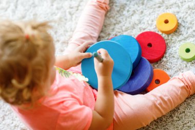 Bebek bir halının üzerinde oturur, renkli ahşap halkalar ve oyuncak bir kamyonla oynar, parçaları istiflemeye odaklanır. Çocuk evde ya da çocuk odasında gökkuşağı renkli piramitlerle oynuyor.