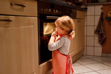 Şirin sarışın kız ev mutfağında kek ya da turta pişiriyor. Mutlu çocuk yardım ederken eğleniyor, Ofen 'in yanında oturuyor ve kurabiyeleri bekliyor. Çocuk Şükran Günü ya da Noel tatili için yemek pişiriyor.
