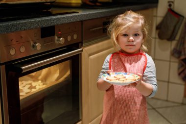 Evcil mutfakta Noel kurabiyesi pişiren sevimli, komik, sarışın bir kız. Çocuk yardım ederken eğleniyor, yanında bir tabak taze pişmiş tatlı kurabiyeyle oturuyor.