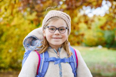 İlkokula giderken küçük şirin bir anaokulu kızının portresi. Okula yürürken gözlük takan sağlıklı, mutlu bir çocuk. Sırt çantalı çocuk, dışarıda. Sarı sonbahar ağaçları.