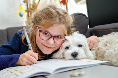 Gözlüklü küçük kız okul ödevini yapıyor. Çalışma kitabında yazan çocuk, öğrenmekle meşgul. Küçük Malta köpeği çocuğun yanında oturuyor, sevimli bir aile hayvanı. İlkokul, eğitim