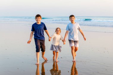 Üç çocuk, gün batımında okyanus sahilinde mutlu kardeşler. Mutlu bir aile, iki okul çocuğu ve küçük bir anaokulu kızı. Kardeşler eğleniyor, İspanya, Costa del Sol