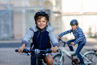 Kasklı iki okul çocuğu şehirde sırt çantalı bisikletle dolaşıyorlar. Renkli giysiler içinde okula bisikletle giden mutlu çocuklar. Çocuklar için güvenli okul yolu..