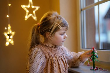 Küçük anaokulu kızı pencerenin yanında oturuyor ve küçük cam Noel ağacını küçük Noel oyuncaklarıyla süslüyor. Mutlu sağlıklı çocuk geleneksel aile tatilini kutluyor.