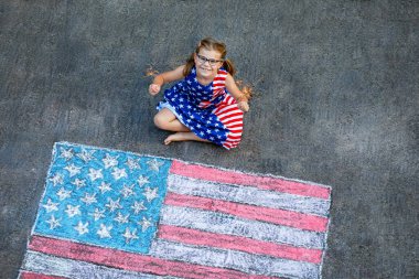 Little girl with glasses and stars and stripes dress, sits on a paved surface beside a large chalk drawing of the American flag. Happy child holding burning sparkler, celebrating holiday in USA clipart