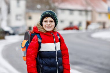 İlköğretim sınıfının küçük çocuğu kar yağarken okula yürüyor. Mutlu çocuk eğleniyor ve ilk karla oynuyor. Gözlüklü öğrenci. Sırt çantasında renkli kış kıyafetleri var.