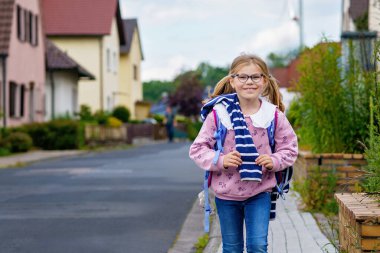 Gözlüklü, pembe kazaklı ve mavi çantalı, ilkokula doğru yürüyen mutlu küçük bir kız. Kendine güvenerek gülümsüyor, güzel bir günde okula gitmeye hazır..