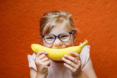 Happy little child girl with yellow banana like smile on orange background. Preschool girl with glasses smiling. Healthy fruits for children.