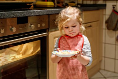 Evcil mutfakta Noel kurabiyesi pişiren sevimli, komik, sarışın bir kız. Çocuk yardım ederken eğleniyor, yanında bir tabak taze pişmiş tatlı kurabiyeyle oturuyor.