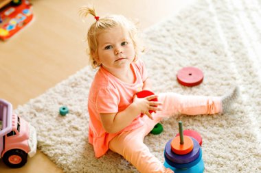 Mutlu küçük kız halıya oturmuş, renkli tahta halkalar ve oyuncak kamyonla oynuyor, parçaları istiflemeye odaklanmış. Çocuk evde ya da çocuk odasında gökkuşağı renkli piramitlerle oynuyor.