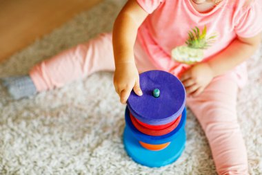 Close-up of toddler sits on a carpet, playing with colorful wooden rings and a toy truck, focused on stacking the pieces. Child plays with rainbow colored pyramid at home or nursery. clipart