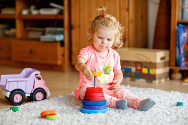 Happy toddler girl sits on a carpet, playing with colorful wooden rings and a toy truck, focused on stacking the pieces. Child plays with rainbow colored pyramid at home or nursery. clipart