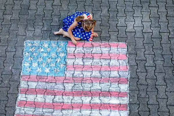 Gözlüklü, yıldız ve çizgili kıyafetler giymiş küçük bir kız, Amerikan bayrağının büyük bir tebeşir çiziminin yanında asfalt bir yüzeye oturur. Mutlu çocuk asfalta Amerikan bayrağı çiziyor.