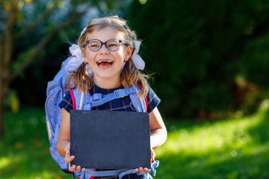 Gözlüklü, sırt çantalı, çantalı ve okulun ilk günü büyük çantalı mutlu bir kız çocuğu. Dışarıda sağlıklı, sevimli bir çocuk var. İlk okul günü için tebeşirleri tutan çocuk