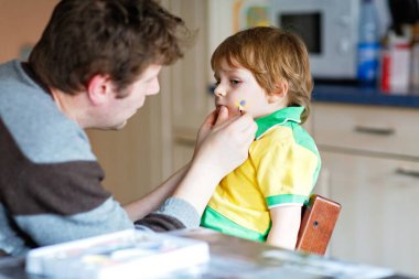 Küçük oğlunun yüzüne futbol ya da futbol maçı için bayrak boyayan genç bir baba. Mutlu anaokulu çocuk hayranı ve babası stadyumda izlemek ve tezahürat yapmak için favori takım maçına hazırlanıyor..