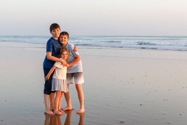 Üç çocuk, gün batımında okyanus sahilinde mutlu kardeşler. Mutlu bir aile, iki okul çocuğu ve küçük bir anaokulu kızı. Kardeşler eğleniyor, İspanya, Costa del Sol