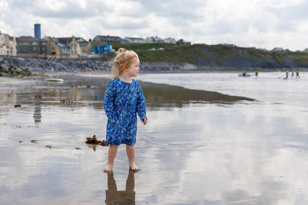 Маленькая милая малышка на пляже Баллибанион, развлекается играя на западном побережье Ирландии. Счастливый ребенок наслаждается ирландским летом и солнечным днем с семьей