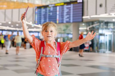 Havaalanı terminalindeki küçük anaokulu kızı. Mutlu çocuk uçakla tatile çıkıyor. Pasaportu ve çantası olan gülümseyen çocuk..