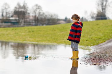 Sarı çizmeli mutlu çocuk baharda ya da sonbahar günü büyük su birikintisinin yanında kağıt gemi teknesiyle oynuyor. Çocuklar için aktif bir eğlence. Komik çocuk dışarıda eğleniyor, renkli kıyafetler giyiyor.