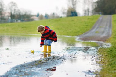 Sarı çizmeli mutlu çocuk baharda ya da sonbahar günü büyük su birikintisinin yanında kağıt gemi teknesiyle oynuyor. Çocuklar için aktif bir eğlence. Komik çocuk dışarıda eğleniyor, renkli kıyafetler giyiyor.