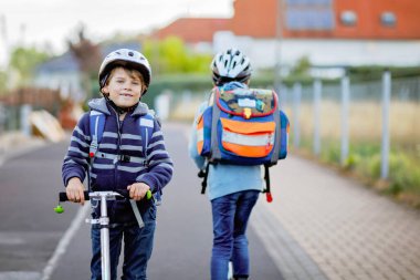 Güneşli bir günde, ellerinde scooterla şehirde gezen kasklı iki okul çocuğu. Renkli giysiler içinde okula giderken bisiklet süren mutlu çocuklar. Çocuklar için güvenli okul yolu..