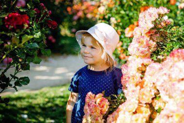 Çiçek açan gül bahçesinde küçük bir kızın portresi. Yaz güneşli bir günde parkta güllerle ve çiçeklerle eğlenen sevimli güzel bir çocuk. Mutlu gülümseyen bebek