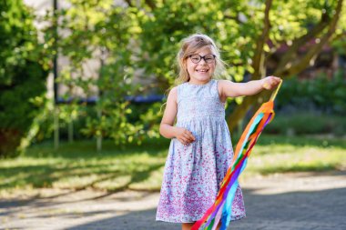 Gözlüklü küçük anaokulu kızı renkli kurdeleyle jimnastik dansı yapıyor. Dışarıda oynayan sevimli bir çocuk. Çocuklar için spor ve etkinlik