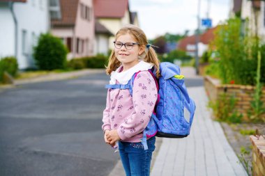 Gözlüklü, pembe kazaklı ve mavi çantalı, ilkokula doğru yürüyen mutlu küçük bir kız. Kendine güvenerek gülümsüyor, güzel bir günde okula gitmeye hazır..