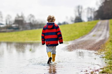 Sarı yağmur giyen küçük çocuk çocuk çizme ve yürüme ve içine atlama bahar günü sıcak güneşli puddle. Eğleniyor ve açık havada oyun renkli moda rahat yağmur giysileri mutlu çocuk