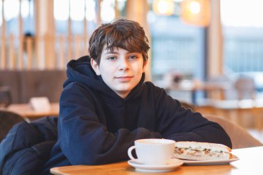 Genç çocuk lokanta kafesinde kahvaltı yapıyor. Mutlu genç çocuk doldurulmuş ekmek yiyip kahve içiyor.