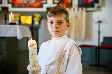 Küçük çocuk ilk kutsal komünyonunu alıyor. Vaftiz mumtutan mutlu çocuk. Katolik curch geleneği. Sunak yakınındaki bir kilisede beyaz geleneksel elbiseli çocuk