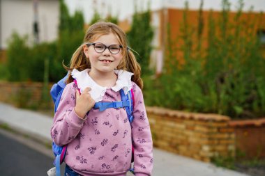 Gözlüklü, pembe kazaklı ve mavi çantalı, ilkokula doğru yürüyen mutlu küçük bir kız. Kendine güvenerek gülümsüyor, güzel bir günde okula gitmeye hazır..