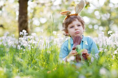 Paskalya tavşanı kulaklı şirin çocuk geleneksel ziyafeti kutluyor. Sıcak ve güneşli bir günde çikolatalı tavşan şekerlemesi yiyen mutlu çocuk. Aile, tatil, bahar konsepti. Çiçeklerin arasında oturan çocuk.