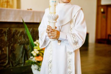 Küçük çocuk ilk kutsal komünyonunu alıyor. Vaftiz mumtutan mutlu çocuk. Katolik curch geleneği. Sunak yakınındaki bir kilisede beyaz geleneksel elbiseli çocuk