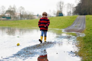 Sarı çizmeli mutlu çocuk baharda ya da sonbahar günü büyük su birikintisinin yanında kağıt gemi teknesiyle oynuyor. Çocuklar için aktif bir eğlence. Komik çocuk dışarıda eğleniyor, renkli kıyafetler giyiyor.