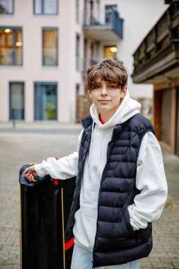 Havalı kıyafetleri, beyaz kapüşonu ve siyah yeleği olan genç bir çocuk büyük bir bavulla dışarıda duruyor, seyahat etmeye hazır.